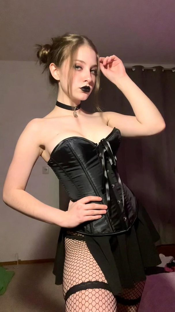 A young woman with brown hair styled in two buns, wearing a black corset, black skirt, and fishnet stockings, posing indoors.