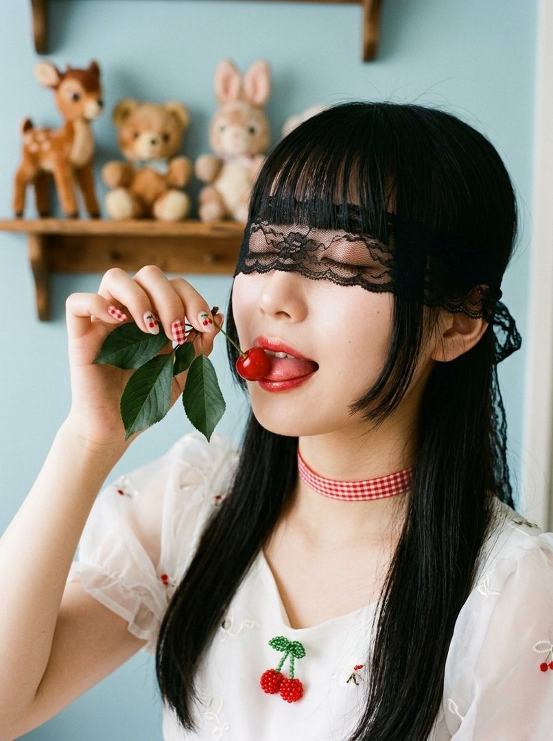 A young woman with black hair and bangs, wearing a white blouse and a red and white checkered choker, is blindfolded with black lace and playfully holds a cherry to her lips.