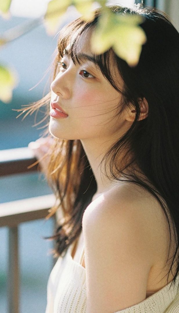 A young East Asian woman with long, dark hair and bangs, looking to the side with a soft expression, bathed in natural light.