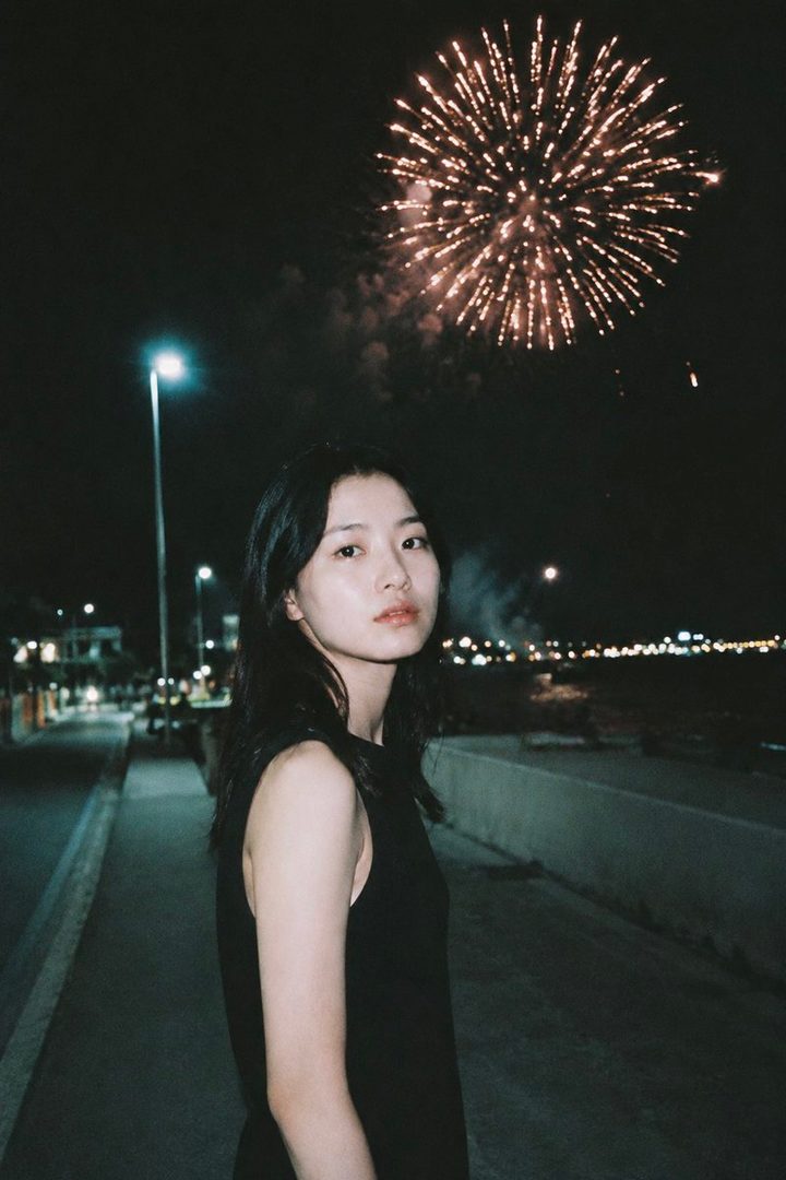 A young woman stands on a street at night with fireworks exploding in the sky behind her.