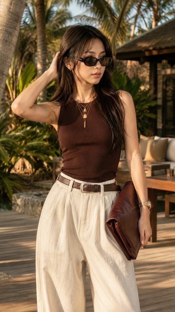 A stylish East Asian woman in a brown ribbed tank top and ivory wide-leg trousers poses confidently in a tropical resort garden during golden hour, holding a maroon clutch bag.