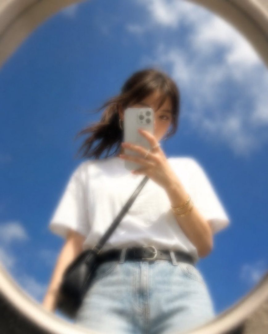 A young woman takes a selfie in a mirror, reflecting a blue sky with clouds. She is wearing a white t-shirt, denim shorts, and a black crossbody bag. The image has a soft, dreamy, and slightly blurred aesthetic.