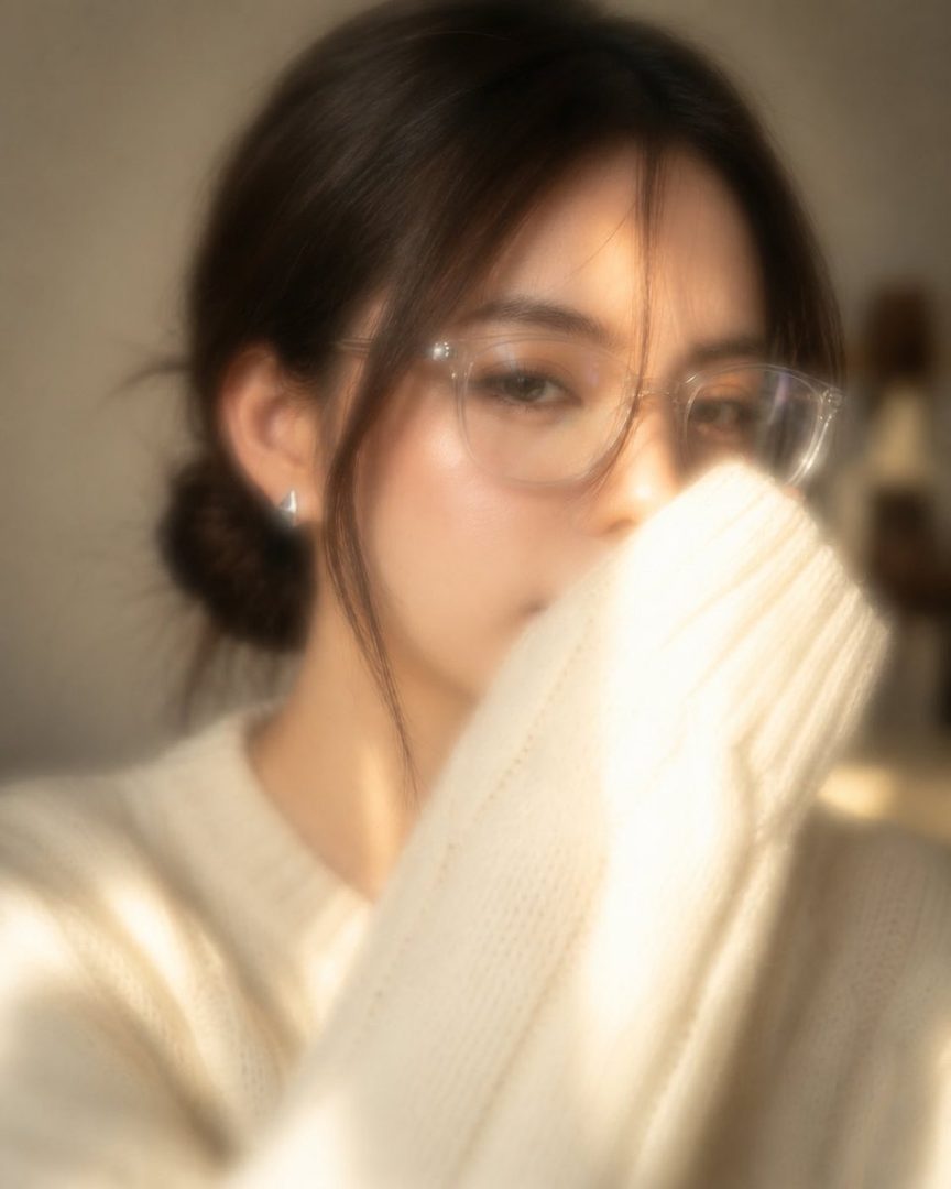 A soft, dreamy portrait of a young East Asian woman with dark hair in a bun, wearing glasses and a cozy knit sweater, partially covering her face with her hand, bathed in warm, diffused light.