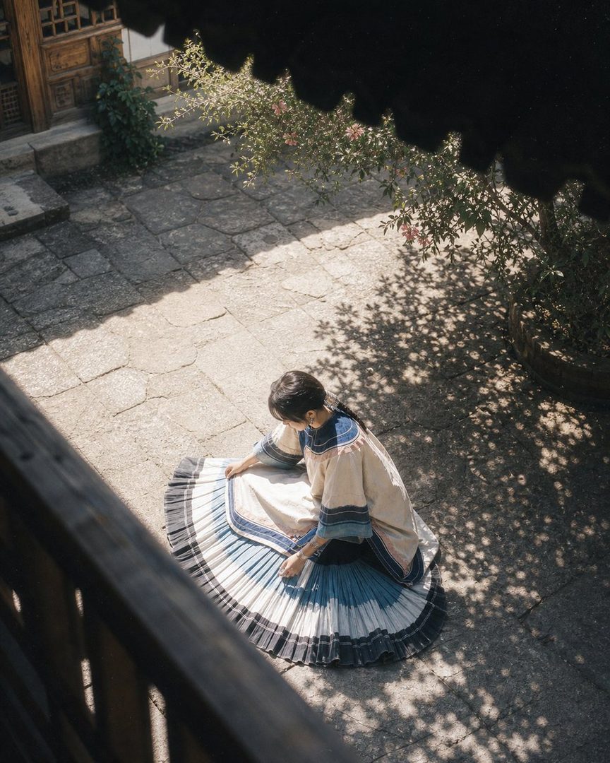 A woman in a traditional dress sits on a stone courtyard, her skirt spread out like a fan, with dappled sunlight and shadows.