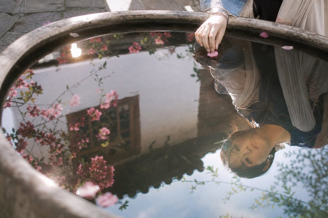 A reflection of a young East Asian woman in a water basin, with pink flowers and a traditional building in the background.