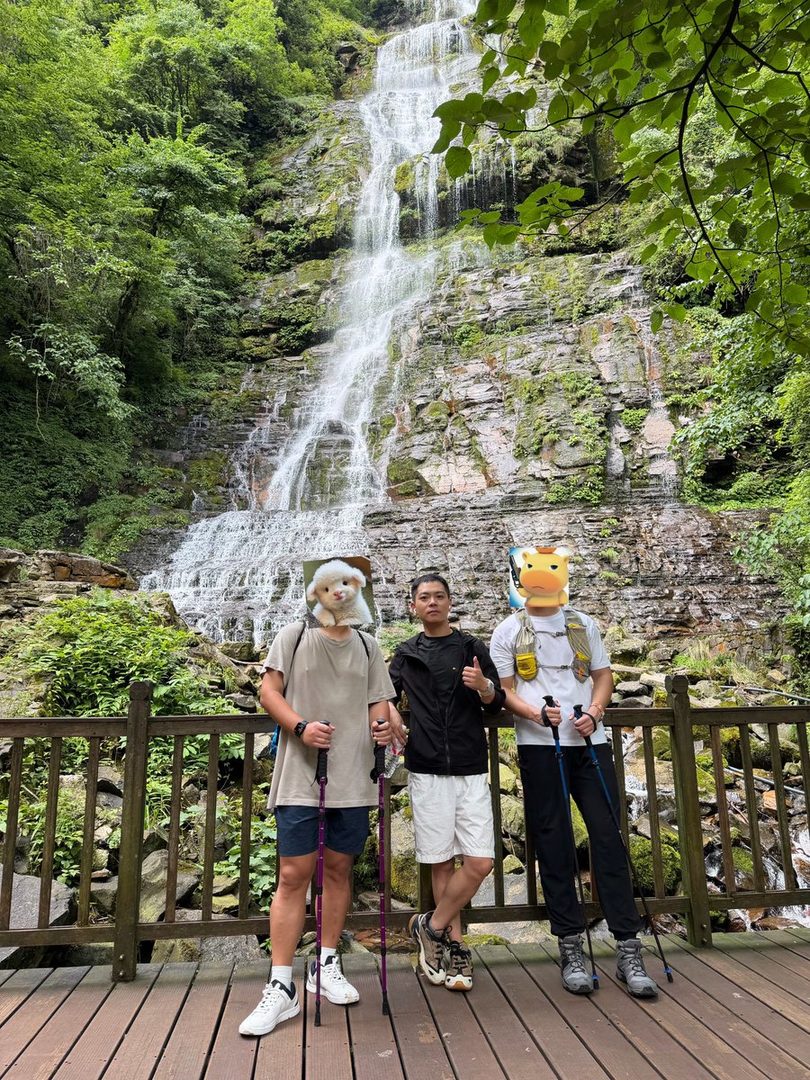 Three people with animal masks stand in front of a waterfall, holding hiking poles.