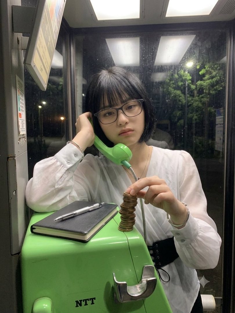 A young woman with short black hair and glasses, wearing a white blouse, is holding a green phone receiver and a stack of coins in a phone booth at night.