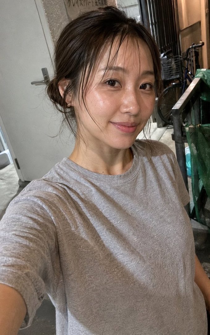 A young East Asian woman with her hair tied up in a messy bun, wearing a grey t-shirt, smiles at the camera in an outdoor setting.