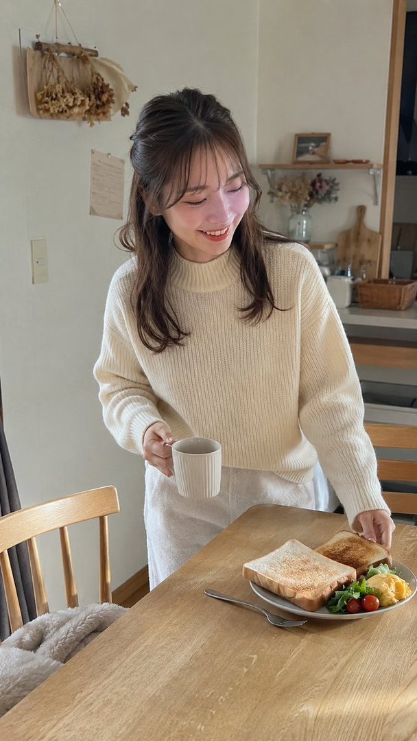 A young East Asian woman in a cream sweater smiles while holding a mug and standing by a table with breakfast.