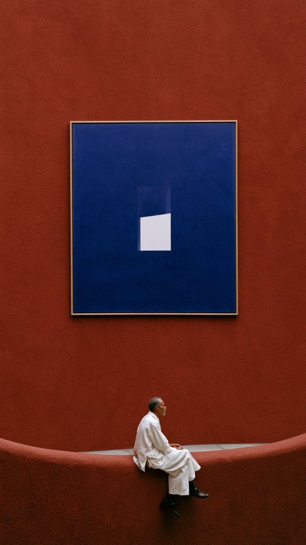 An elderly East Asian man in a white robe sits on a curved ledge in front of a large blue abstract painting on a red wall.