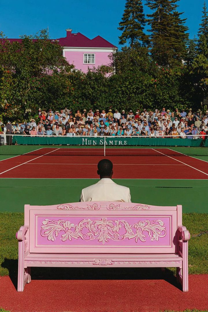 A man in a white suit sits on a pink bench facing a tennis court with spectators in the background.