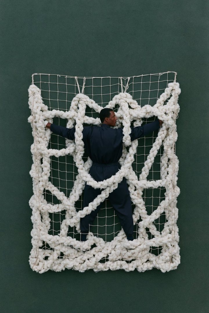 A man in a dark blue jumpsuit is entangled in a large, white, rope-like sculpture against a dark green background.