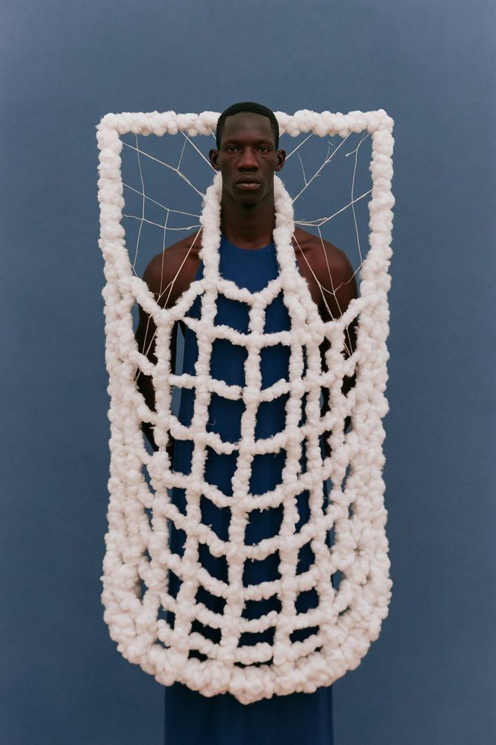 A man wearing a blue tank top and a large, white, net-like costume stands against a blue background.
