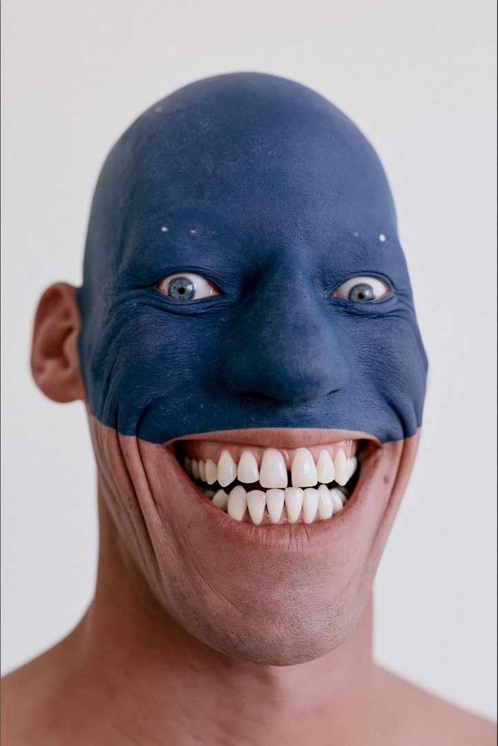 A close-up portrait of a person with their face painted blue, revealing a wide, unnatural smile with prominent teeth.