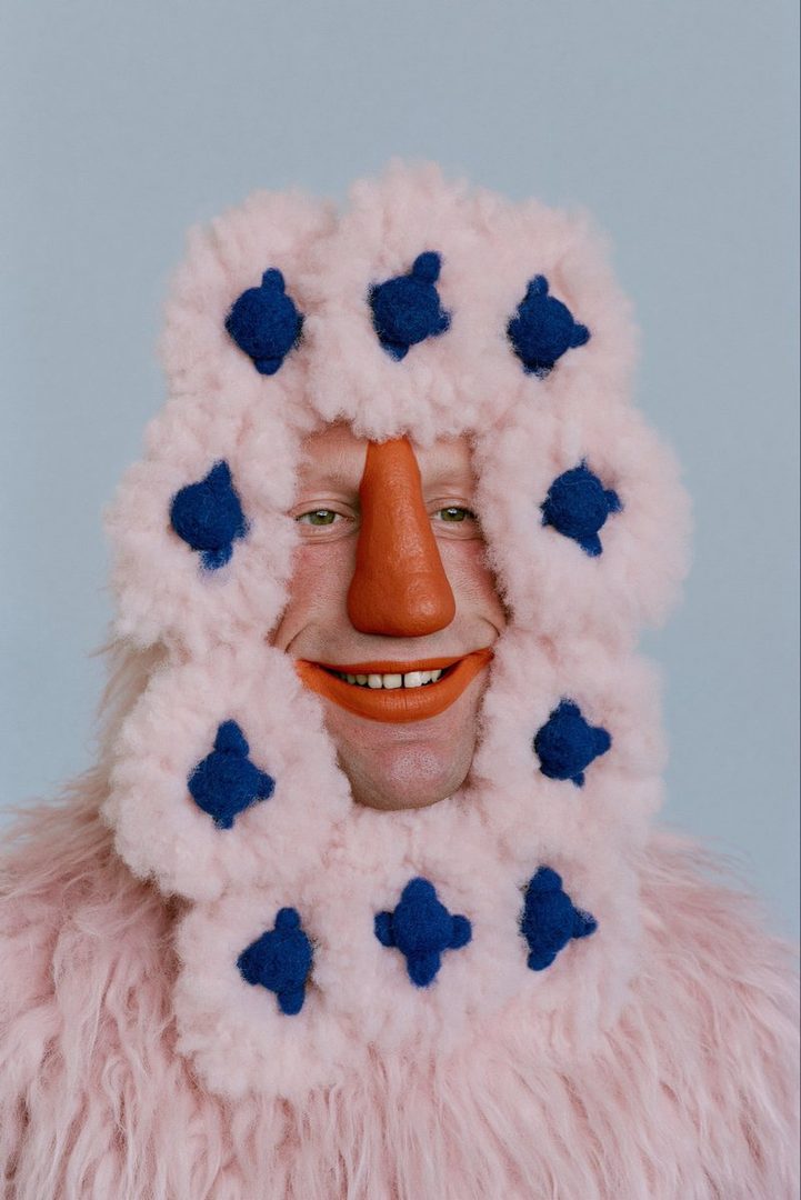 A person wearing a fluffy pink costume with blue accents and an orange nose and lips, smiling against a light blue background.