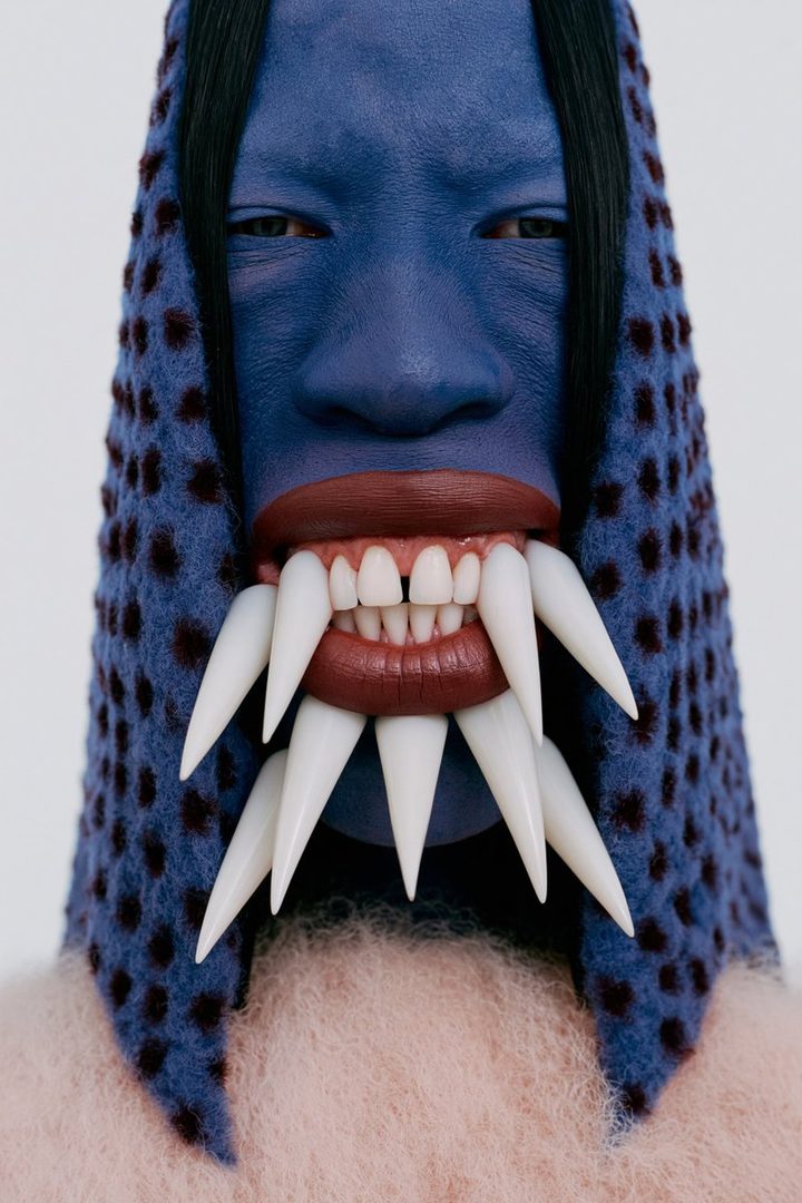 A person with blue face paint and exaggerated fangs, wearing a patterned hood and fluffy sweater.