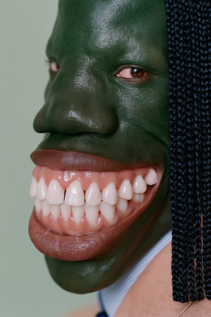 A close-up portrait of a person with green-painted skin, wearing a suit and showing an exaggerated set of white teeth.