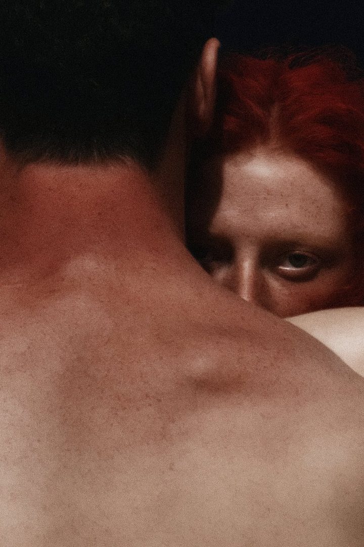 A close-up portrait of a woman with red hair peeking from behind a man's shoulder, with a dark background.