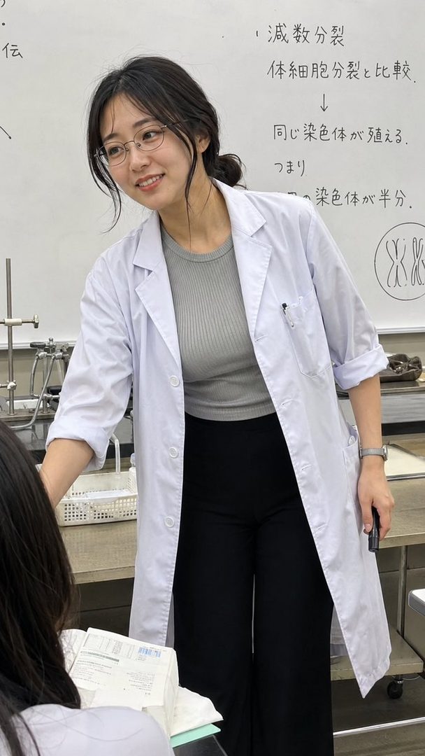 A young female teacher in a lab coat smiles while standing in front of a whiteboard with biological diagrams and text.