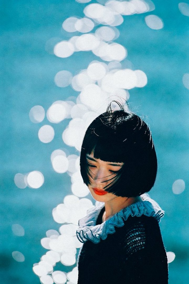 A young woman with short black hair and bangs, wearing a dark sweater with a ruffled collar, looks down with her eyes closed, against a backdrop of shimmering blue water.