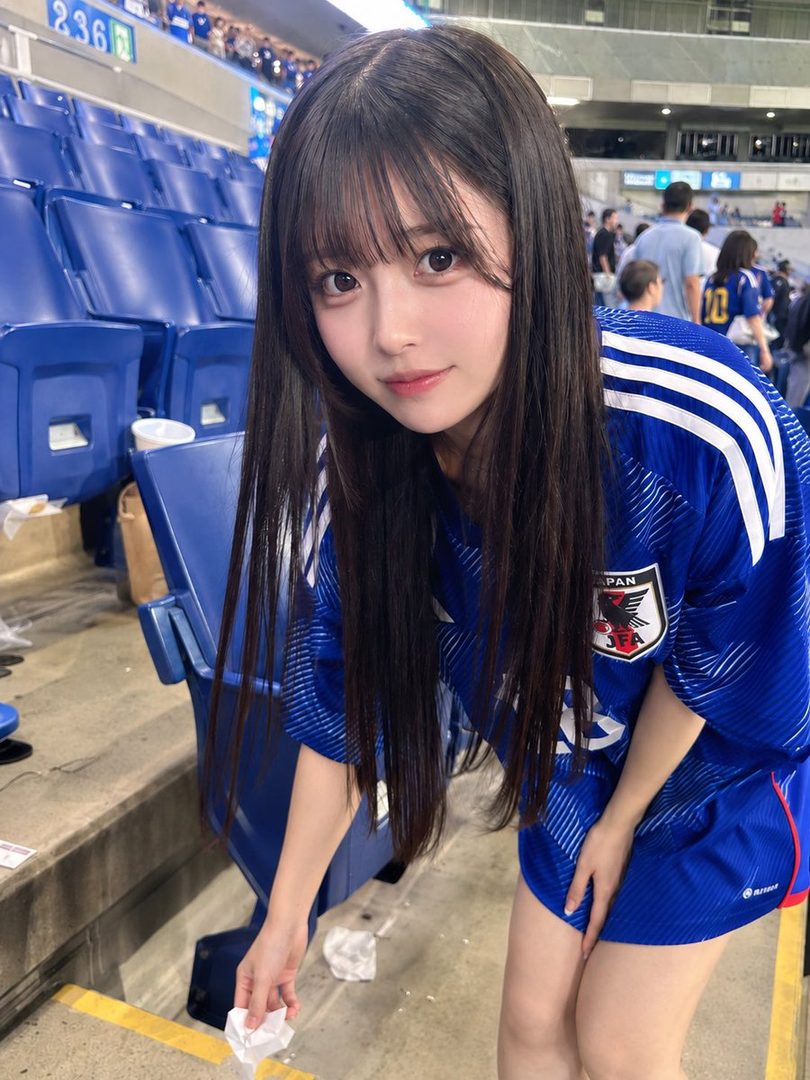 A young woman in a Japanese soccer jersey smiles at the camera while sitting in stadium seats.