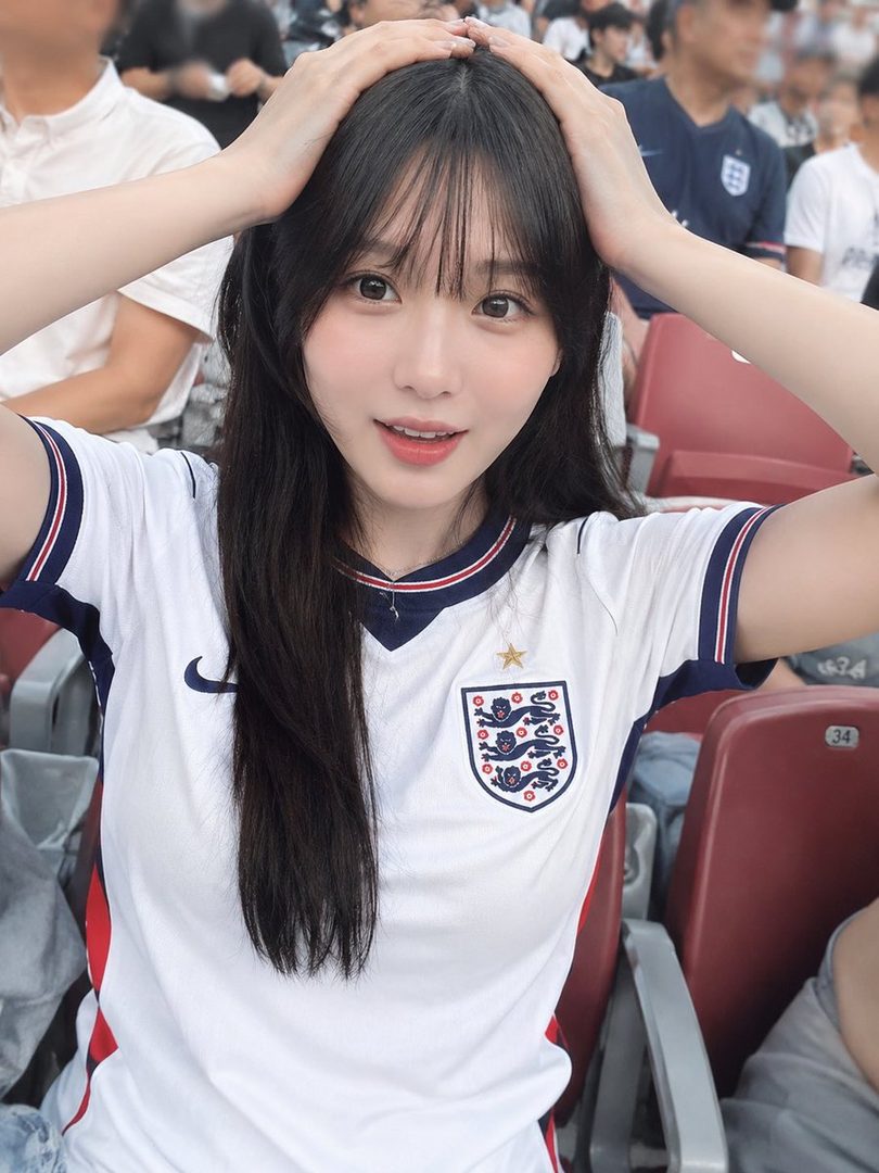 A young woman with long black hair and bangs, wearing an England national football team jersey, poses with her hands on her head in a stadium.