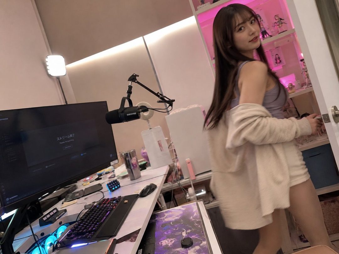 A young woman with long brown hair, wearing a light purple tank top and a beige cardigan, poses playfully in a room with a computer setup and shelves filled with figurines.