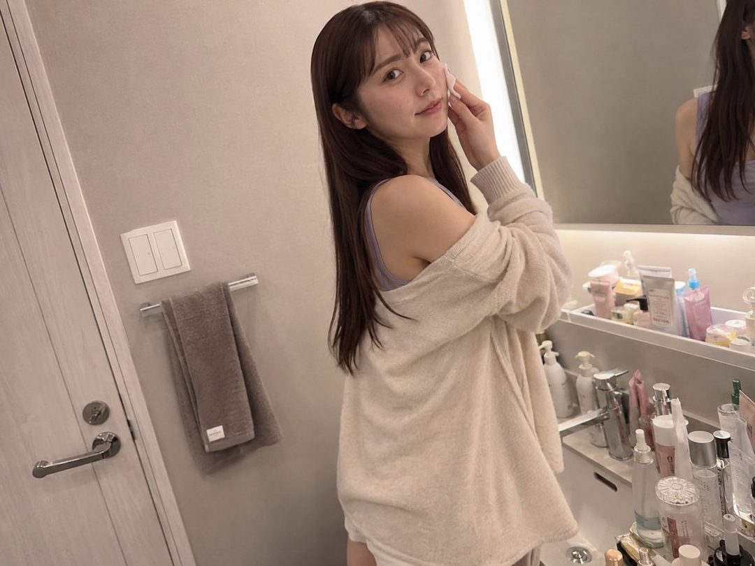 A young East Asian woman with long brown hair is taking a selfie in a bathroom, applying skincare with a cotton pad.