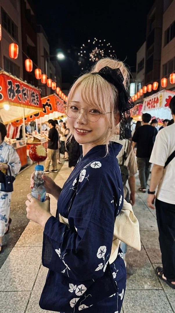 A young woman with blonde hair tied up, wearing glasses and a dark blue yukata with white floral patterns, smiles while holding a candy apple and a bottle of drink at a summer night festival with fireworks in the background.