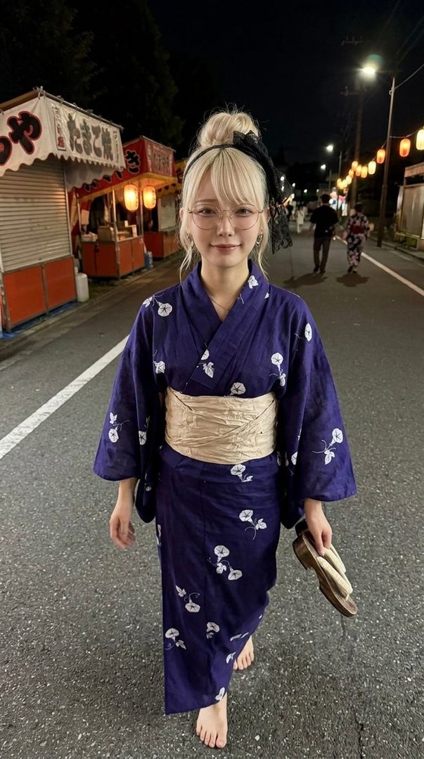 A young woman with blonde hair tied up in a bun, wearing glasses and a blue yukata with white floral patterns, walks barefoot down a street at night, holding a pair of geta sandals.