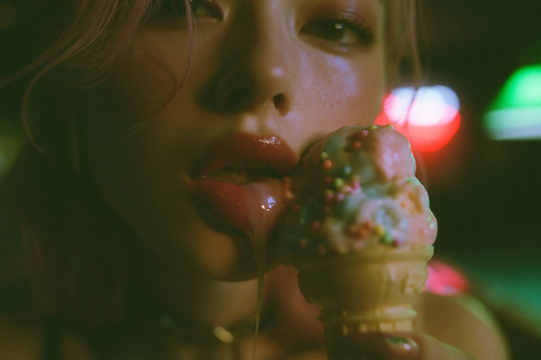 Close-up portrait of a woman with pink hair licking ice cream, with colorful bokeh lights in the background.