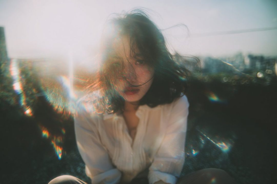 A dreamy portrait of a young East Asian woman with dark hair, wearing a white blouse, sitting outdoors with a city skyline in the background and lens flare effects.