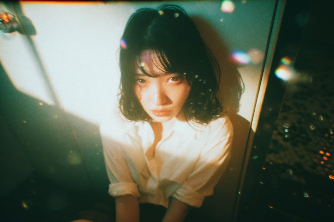 A young woman with dark, wavy hair and bangs, wearing a white button-up shirt, looks directly at the camera with a melancholic expression. The image has a dreamy, cinematic quality with soft lighting and bokeh effects.