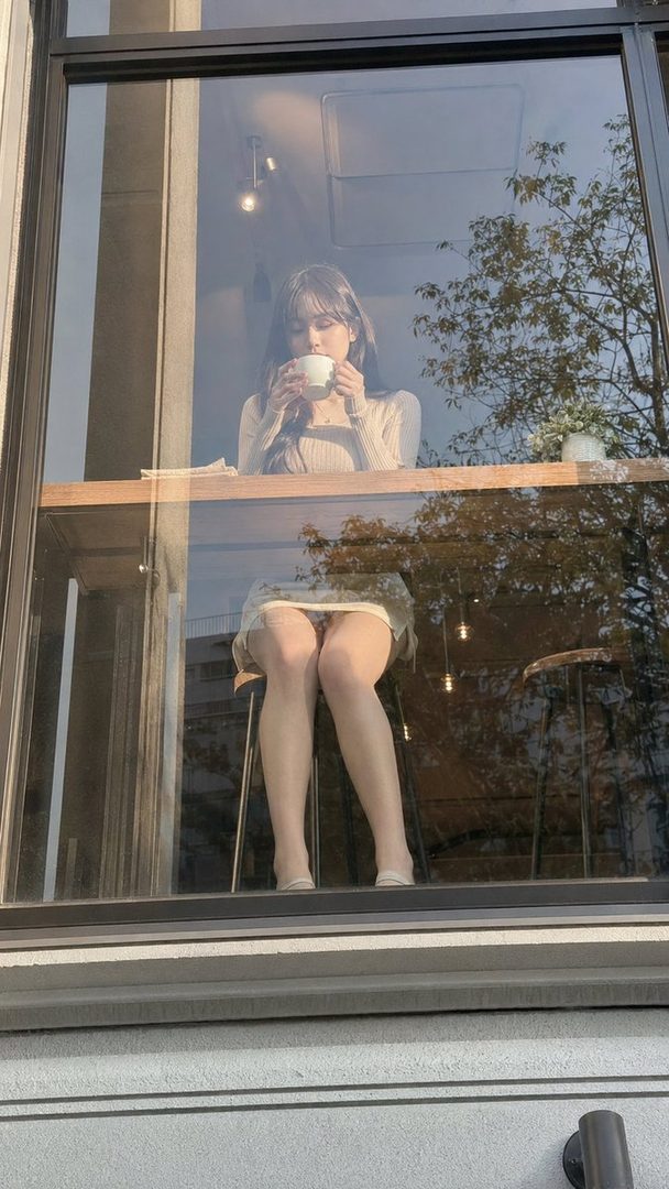 A young woman with long dark hair sits at a cafe window, holding a cup of coffee and looking down.