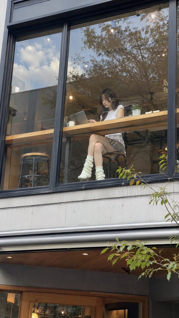 A young woman with long dark hair sits at a table in a cafe, looking at her laptop.