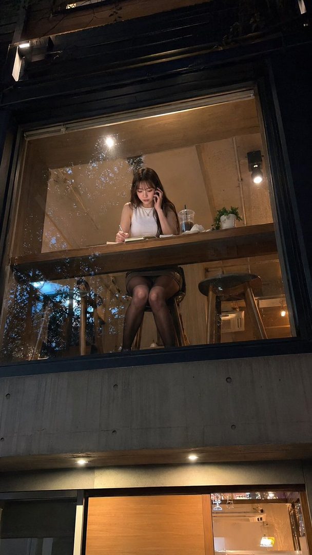 A young woman with long brown hair sits at a table in a cafe at night, writing in a notebook and talking on the phone.