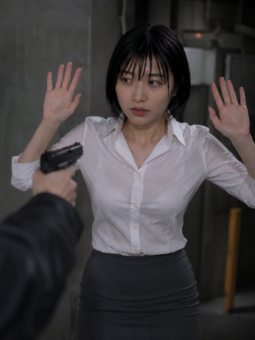 A woman in a white blouse and grey skirt holds her hands up in surrender as a gun is pointed at her.