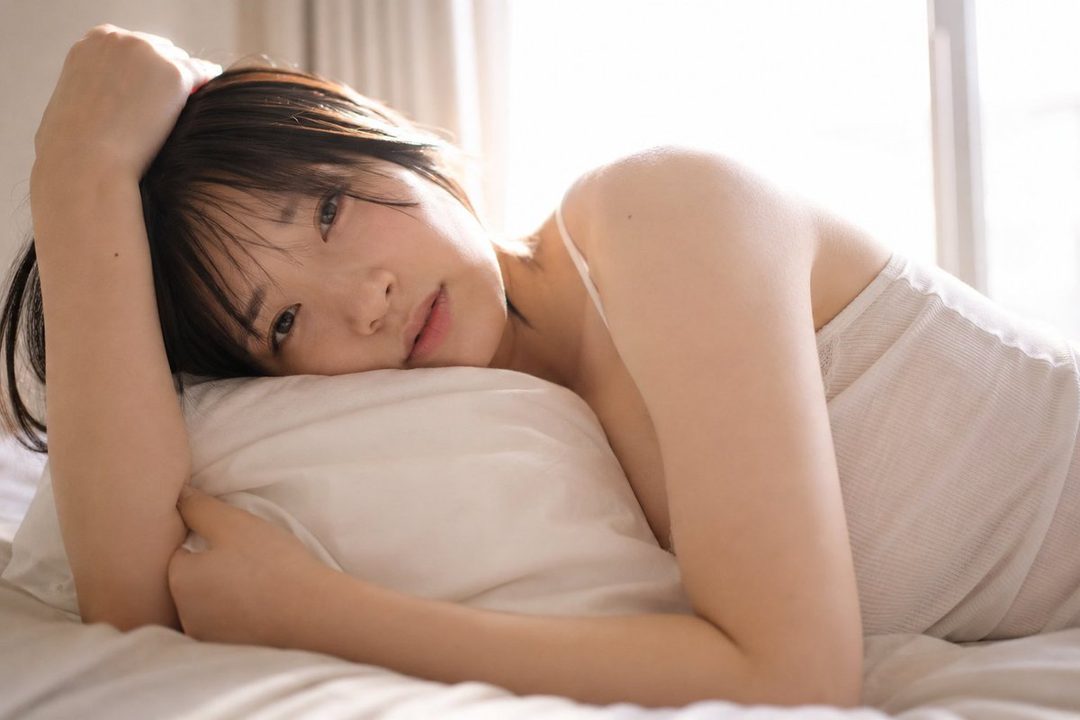 A young woman with short black hair lies on a pillow, looking towards the camera with a soft expression. Natural light streams in from the side, creating a dreamy atmosphere.
