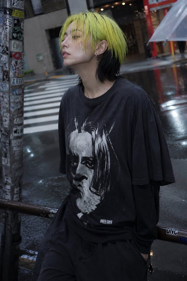 A person with bright yellow and black hair, wearing a dark t-shirt with a graphic print, stands on a wet city street.
