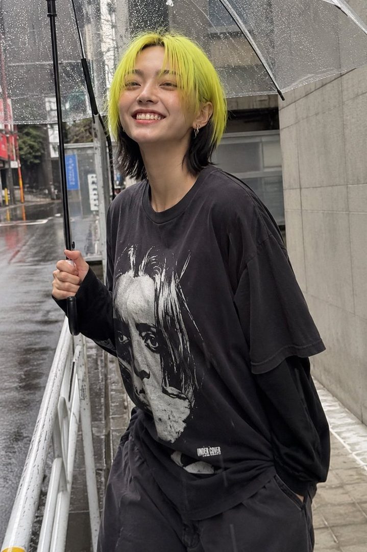 A young woman with bright yellow and black hair smiles while holding a clear umbrella on a rainy street.