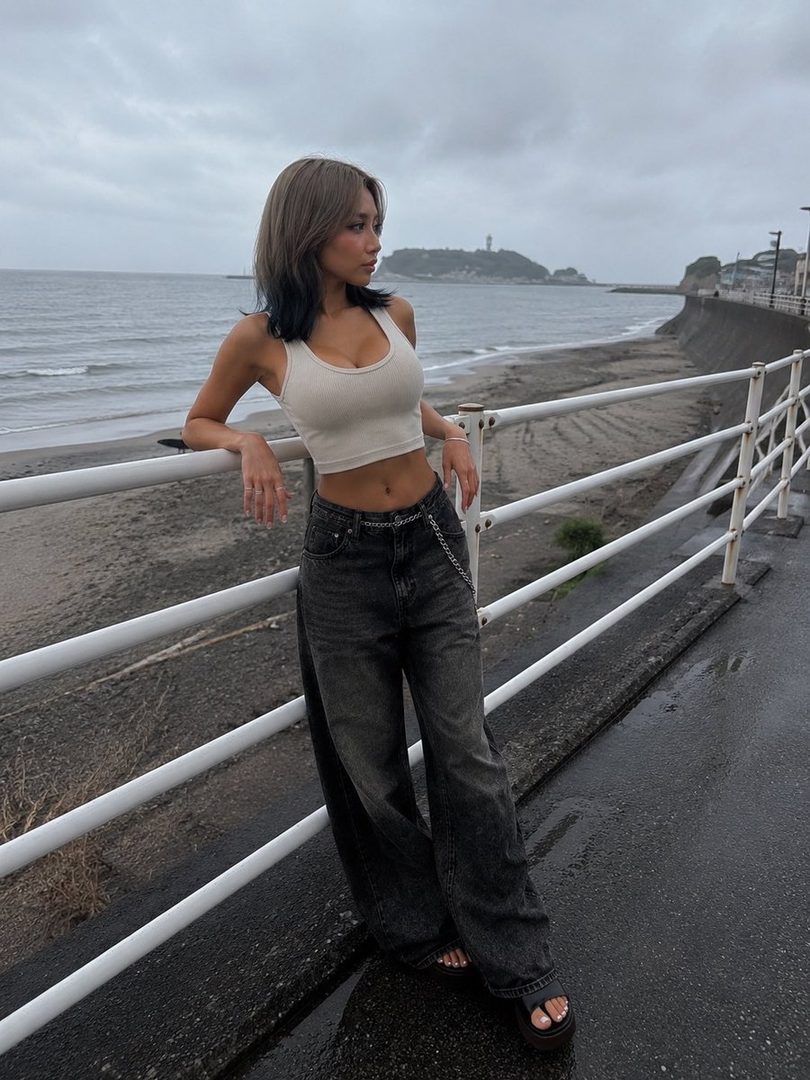 A young woman with shoulder-length brown and black ombre hair, wearing a white crop top and dark wide-leg jeans, leans against a railing by the sea on an overcast day.