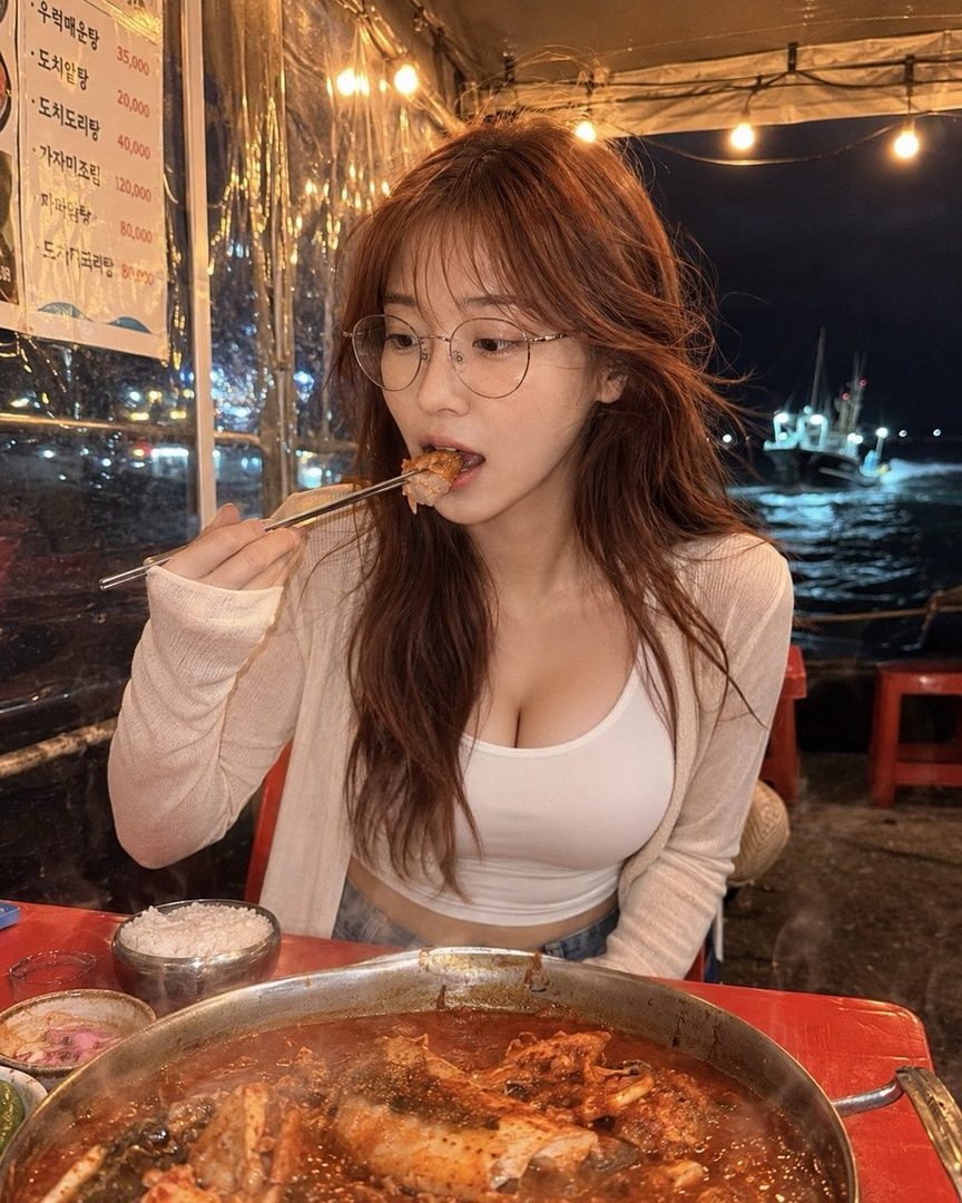 A young woman with long brown hair and glasses eating a meal at a seaside restaurant at night.