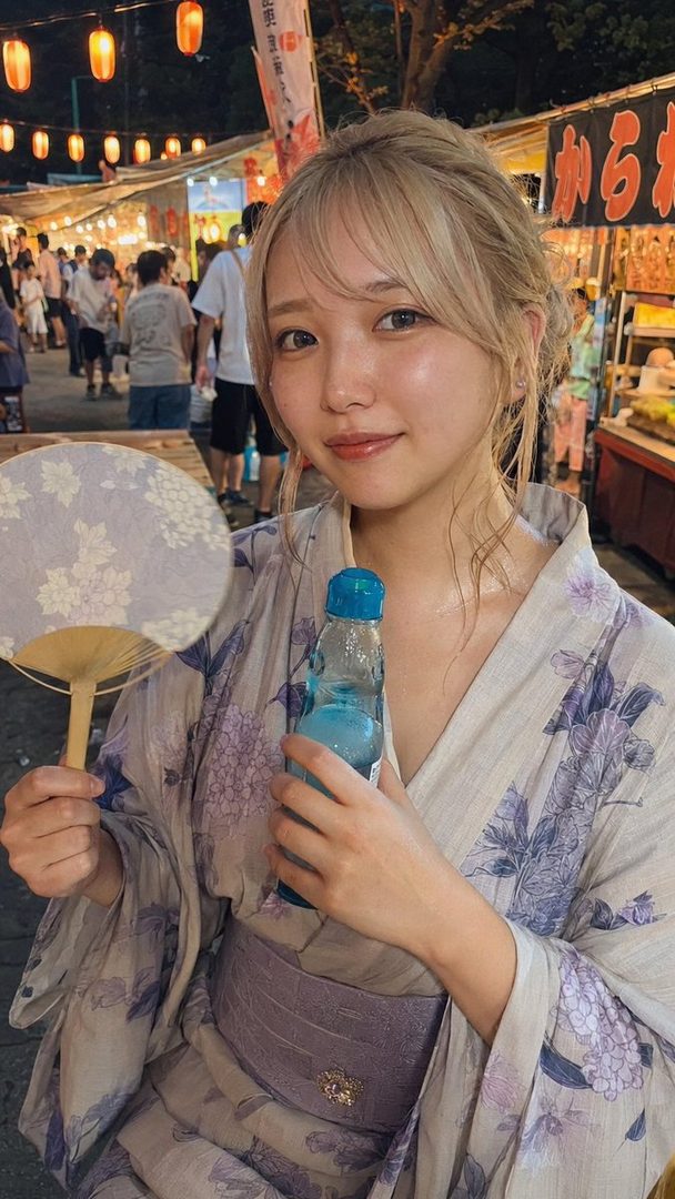 A young Japanese woman with blonde hair, wearing a floral yukata and holding a traditional fan and a bottle of Ramune soda, at a summer festival at night.