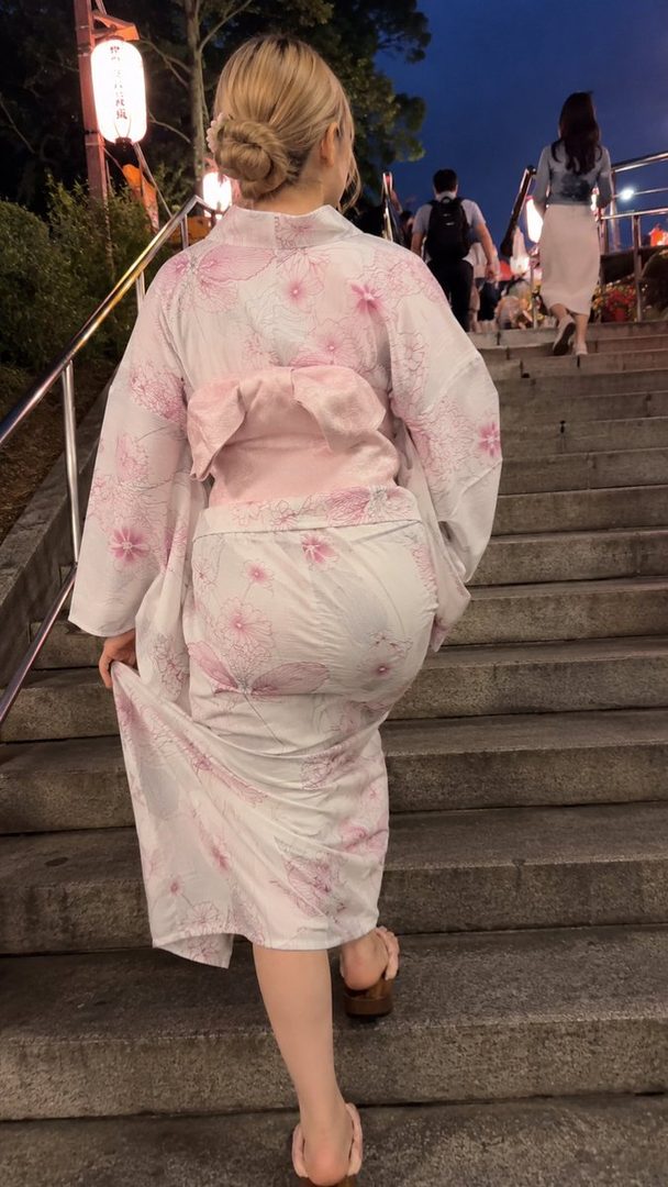 A woman with blonde hair tied in a bun, wearing a pink and white floral yukata, walks up stone stairs at night, illuminated by lanterns.