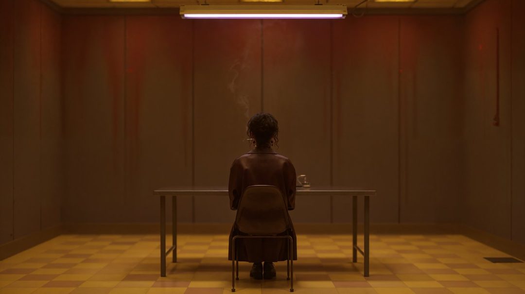 A person sits alone at a table in a dimly lit room, with smoke rising from a cigarette.