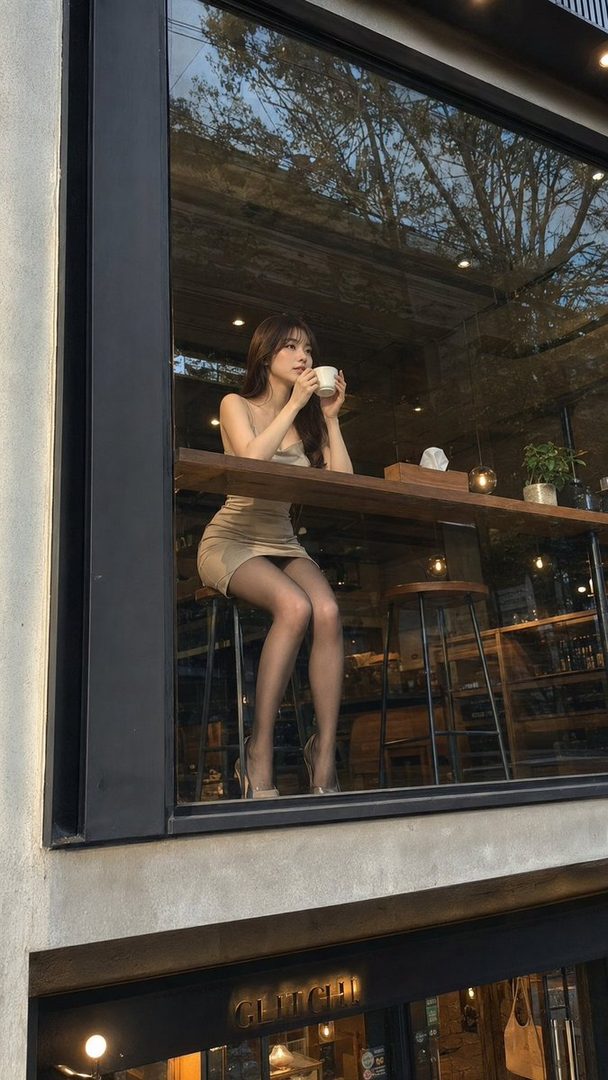 A young East Asian woman in a tight dress and stockings sits at a cafe counter, holding a coffee cup.