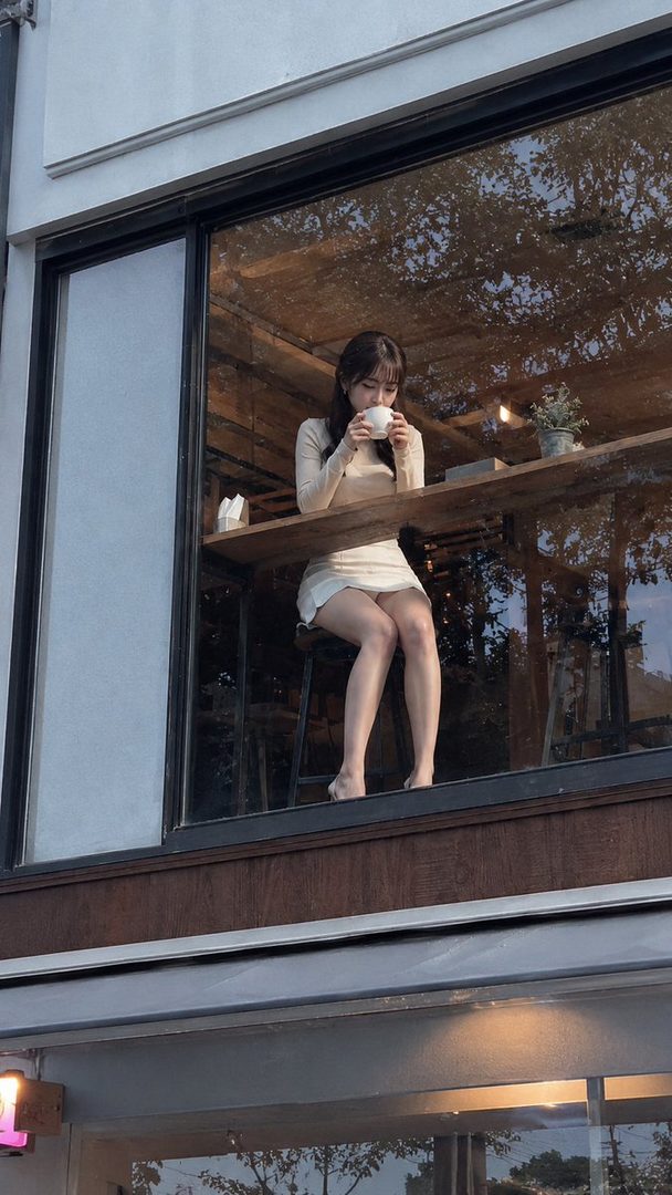 A young woman with long dark hair sits at a cafe counter, drinking from a white mug.