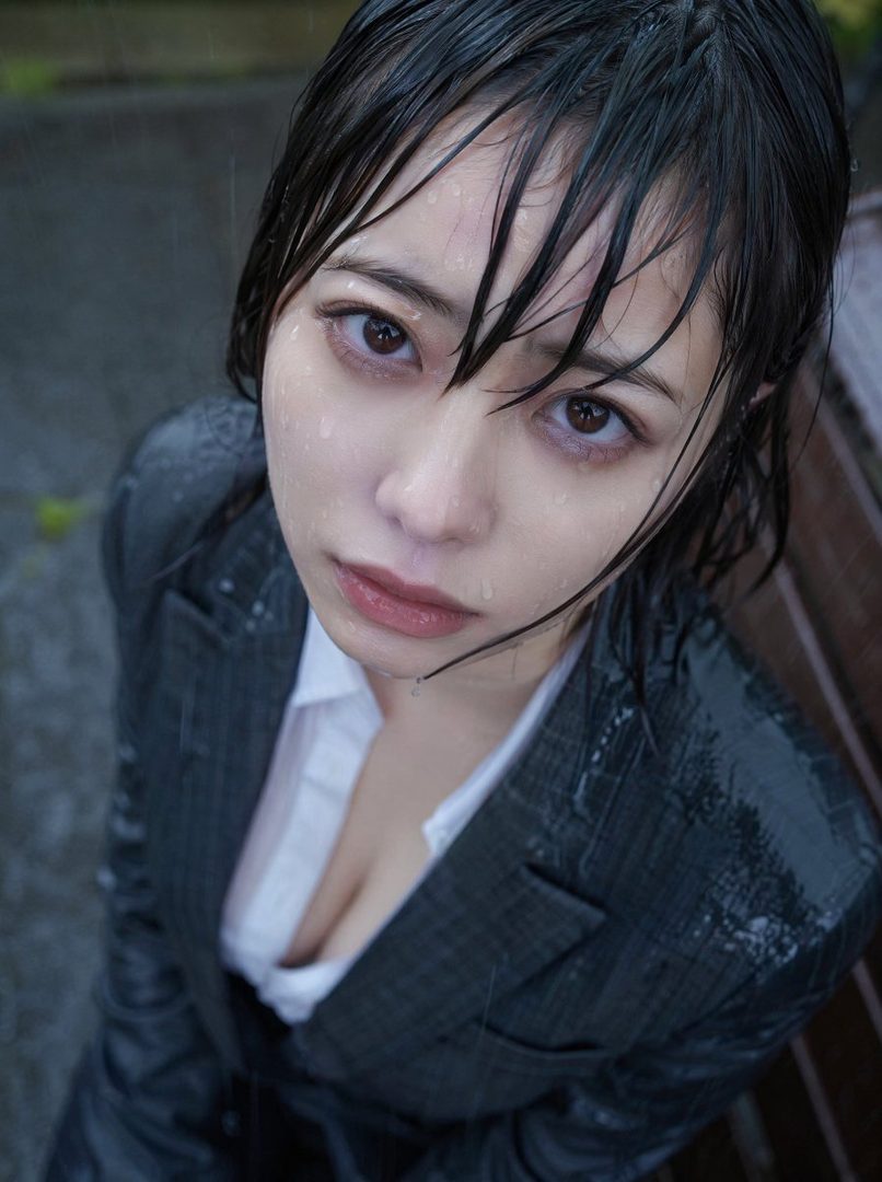 A woman in a wet suit jacket and white shirt looks up in the rain.