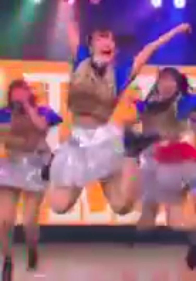 Three young women in sparkly skirts and tops jumping on a stage with colorful lighting.