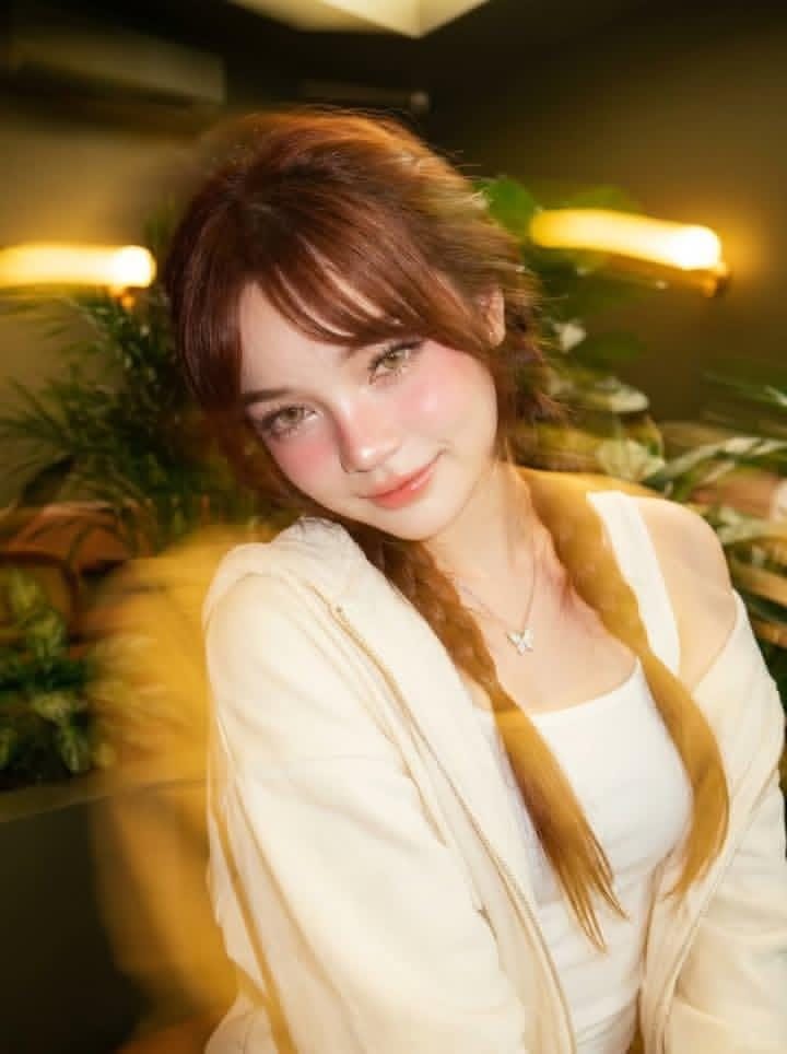 A photorealistic portrait of a young woman with brown braided hair, wearing a white tank top and an off-white hoodie, sitting indoors with plants in the background.